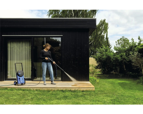 Femme nettoie terrasse en bois avec nettoyeur haute pression à côté d'une maison noire.