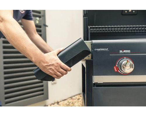 Un homme insère une batterie noire dans un barbecue à gaz Campingaz.