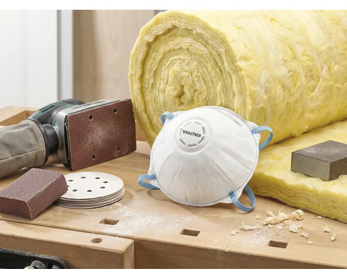 Scène avec masque antipoussière, papier abrasif, ponceuse et laine d'isolation sur un établi.