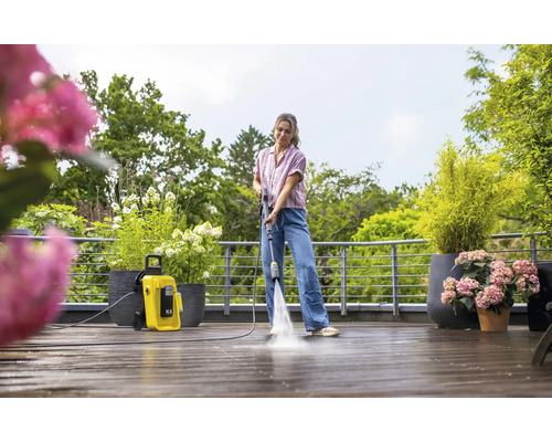 Eine Frau reinigt eine Holzterrasse mit einem Kärcher Hochdruckreiniger K4. Pflanzen und Bäume sind im Hintergrund zu sehen.