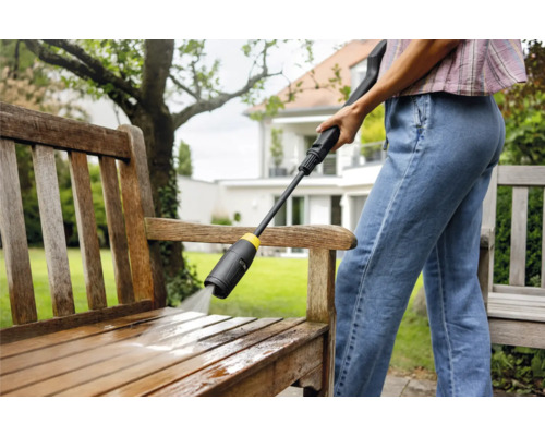 Frau reinigt eine Holzbank mit einem Hochdruckreiniger im Garten.