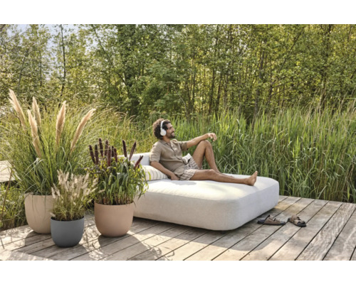 Scène de jardin avec un homme allongé sur un canapé de salon, entouré d'herbes ornementales dans des jardinières.