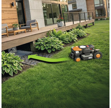 Un robot tondeuse à gazon se dirige sur un espace vert vers sa station de charge sous une terrasse en bois.