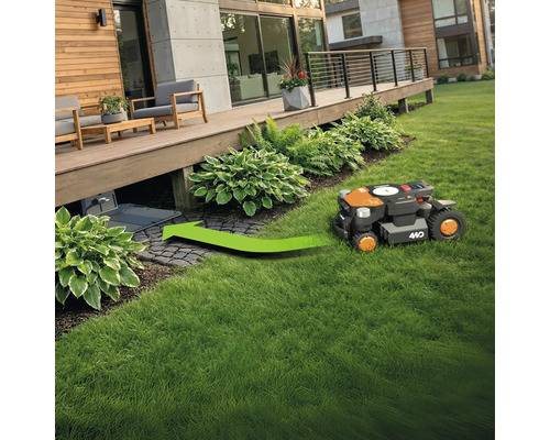 Un robot tondeuse à gazon se dirige sur un espace vert vers sa station de charge sous une terrasse en bois.