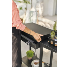 Eine Person bereitet eine Grillplatte auf einem schwarzen Grilltisch vor, der mit einem Drehknopf und einem Behälter ausgestattet ist. Auf dem Tisch stehen Kräuter und Gewürze.