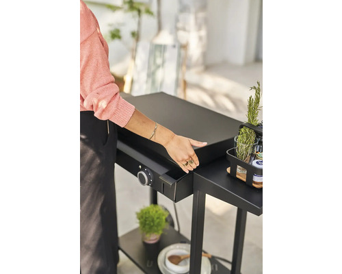 Eine Person bereitet eine Grillplatte auf einem schwarzen Grilltisch vor, der mit einem Drehknopf und einem Behälter ausgestattet ist. Auf dem Tisch stehen Kräuter und Gewürze.