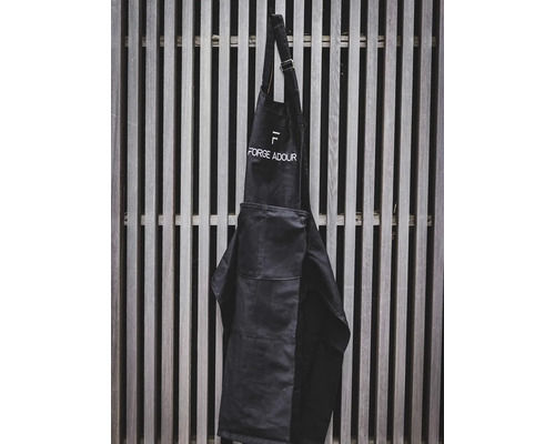 Schwarze Werkstattschürze mit aufgesetzter Tasche und Logo hängt an einer Holzlattenwand.