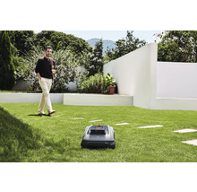 Un robot tondeuse à gazon sur une pelouse, avec un homme se promenant avec une boisson en arrière-plan.