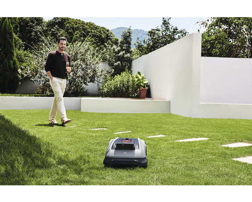 Un robot tondeuse à gazon sur une pelouse, avec un homme se promenant avec une boisson en arrière-plan.