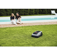 Robot tondeuse sur pelouse devant un couple assis au bord d'une piscine.