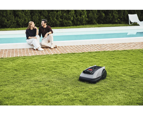 Robot tondeuse sur pelouse devant un couple assis au bord d'une piscine.