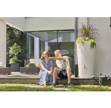 Ein Mann und eine Frau sitzen auf einer modernen Terrasse neben einer hohen weissen Regentonne mit strukturierter Oberfläche und Pflanzschale.