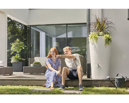 Ein Mann und eine Frau sitzen auf einer modernen Terrasse neben einer hohen weissen Regentonne mit strukturierter Oberfläche und Pflanzschale.