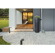 Moderner Regenspeicher mit integrierter Pflanzschale auf einer gepflasterten Terrasse neben einer Hauswand und einer Gießkanne aus Metall.