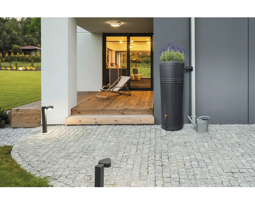 Moderner Regenspeicher mit integrierter Pflanzschale auf einer gepflasterten Terrasse neben einer Hauswand und einer Gießkanne aus Metall.