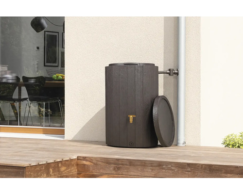 Regentonne in Holzoptik mit goldenem Wasserhahn und Deckel auf einer Holzterrasse an einer Hauswand, angeschlossen an ein Fallrohr.