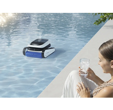 Une piscine avec un robot de nettoyage de piscine dans l'eau et une femme avec un verre d'eau au bord de la piscine.
