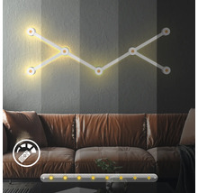 Modulare LED Wandleuchte mit Dimmfunktion über einem Sofa im Wohnzimmer inklusive Symbolen für Fernbedienung und Helligkeitssteuerung.