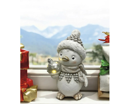 Figurine de Noël pingouin avec bonnet, écharpe et boule de Noël lumineuse sur un rebord de fenêtre devant un paysage d hiver.