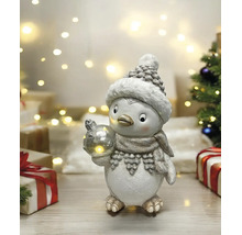 Figurine de Noël pingouin avec bonnet et écharpe tenant une boule lumineuse dans une scène lumineuse avec des cadeaux et des lumières de fête.