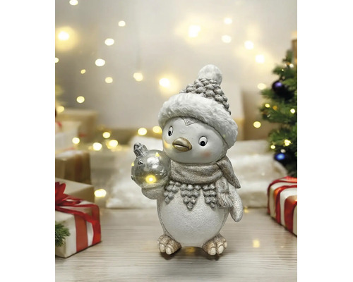 Figurine de Noël pingouin avec bonnet et écharpe tenant une boule lumineuse dans une scène lumineuse avec des cadeaux et des lumières de fête.