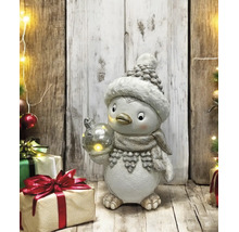 Figurine de pingouin avec bonnet, écharpe et boule lumineuse dans une scène de Noël avec cadeaux devant un mur en bois rustique.