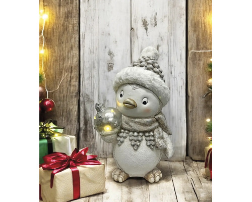 Figurine de pingouin avec bonnet, écharpe et boule lumineuse dans une scène de Noël avec cadeaux devant un mur en bois rustique.