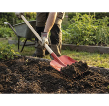 Eine Person gräbt mit einem Spaten im Garten die Erde um. Eine Schubkarre steht im Hintergrund.