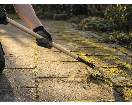 Eine Person entfernt mit einem Fugenreiniger Moos zwischen Pflastersteinen.
