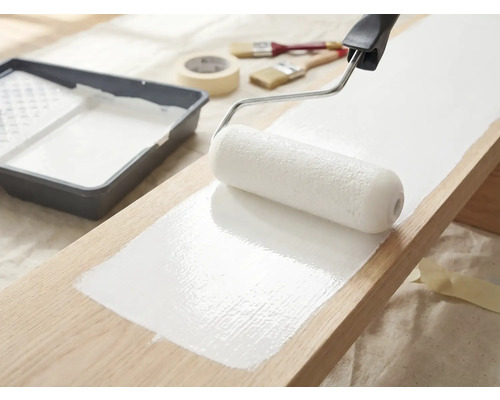 Une planche en bois est peinte avec un rouleau à peinture et de la peinture blanche, un bac à peinture, des pinceaux et du ruban de masquage sont prêts.