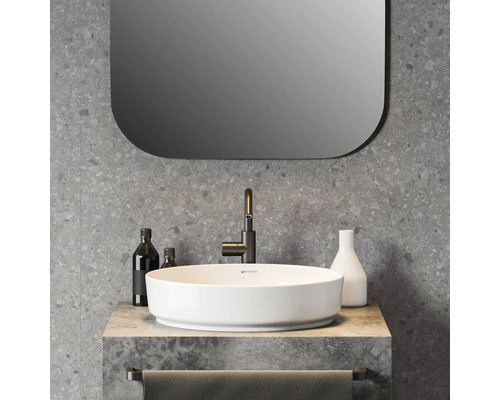 Salle de bains moderne avec vasque à poser ronde blanche, robinetterie noire, mur aspect terrazzo et miroir aux coins arrondis.