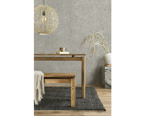 Papier peint intissé aspect pierre en gris, associé à une table en bois, un banc en bois, un tapis tissé foncé et une suspension en osier.