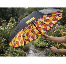 Regenschirm in einem regnerischen Garten mit schwarzer Aussenseite, FloraSelf Logo und buntem Blumenmuster auf der Innenseite.