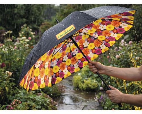Regenschirm in einem regnerischen Garten mit schwarzer Aussenseite, FloraSelf Logo und buntem Blumenmuster auf der Innenseite.