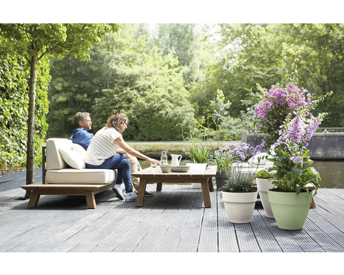 Terrasse de jardin lumineuse avec terrasse en bois, canapé lounge avec coussins, table en bois et diverses plantes en pots au bord de l'eau.