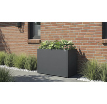 Jardinière rectangulaire gris foncé avec des plantes vertes et des fleurs roses contre un mur en briques sur une terrasse avec des herbes.