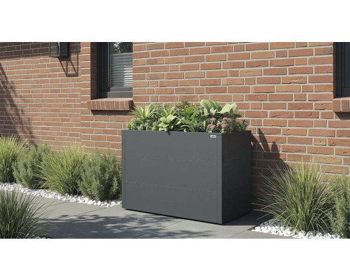 Jardinière rectangulaire gris foncé avec des plantes vertes et des fleurs roses contre un mur en briques sur une terrasse avec des herbes.