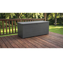 Coffre de jardin en métal gris foncé sur une terrasse en bois avec balustrade devant un jardin.