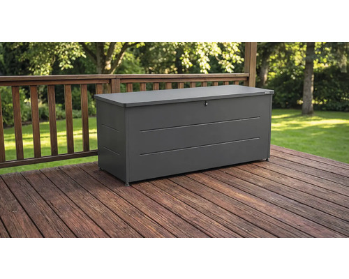 Coffre de jardin en métal gris foncé sur une terrasse en bois avec balustrade devant un jardin.