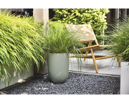 Ziergras in einem hohen, strukturierten grünen Blumentopf auf einer modernen Terrasse mit grauem Splitt und einem hölzernen Liegestuhl.