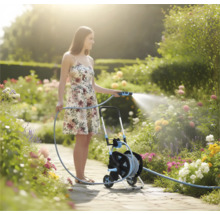 Frau bewässert einen blühenden Garten mit einem Gartenschlauch auf einem Schlauchwagen auf einem hellen Gartenweg.