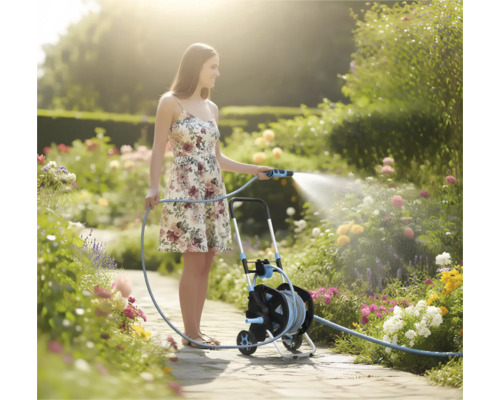 Frau bewässert einen blühenden Garten mit einem Gartenschlauch auf einem Schlauchwagen auf einem hellen Gartenweg.