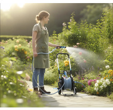 Eine Frau in einer hellen Gartenanlage bewässert Blumen mit einem Gartenschlauch von einem mobilen Schlauchwagen auf einem gepflasterten Weg.