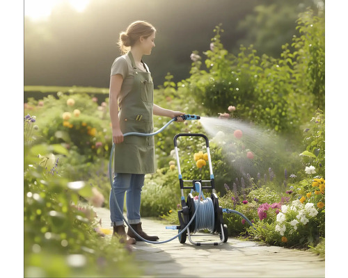 Eine Frau in einer hellen Gartenanlage bewässert Blumen mit einem Gartenschlauch von einem mobilen Schlauchwagen auf einem gepflasterten Weg.