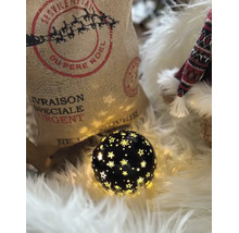 Une boule décorative noire lumineuse avec découpes en étoile sur fausse fourrure blanche à côté du sac de Noël en jute et une figurine.