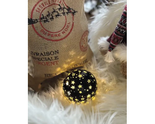 Une boule décorative noire lumineuse avec découpes en étoile sur fausse fourrure blanche à côté du sac de Noël en jute et une figurine.