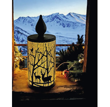 Bougie décorative à LED illuminée avec silhouette de forêt et de cerfs sur un rebord de fenêtre devant un paysage de montagne enneigé.