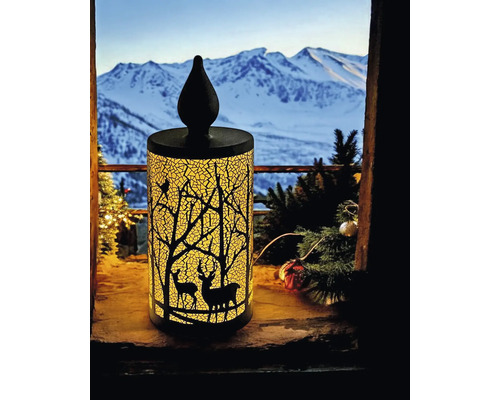 Bougie décorative à LED illuminée avec silhouette de forêt et de cerfs sur un rebord de fenêtre devant un paysage de montagne enneigé.