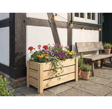 Rechteckiger Blumenkasten aus hellem Holz, bepflanzt mit bunten Blumen, vor einer Fachwerkwand auf einer Steinterrasse neben einer Holzbank.