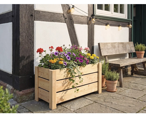 Rechteckiger Blumenkasten aus hellem Holz, bepflanzt mit bunten Blumen, vor einer Fachwerkwand auf einer Steinterrasse neben einer Holzbank.